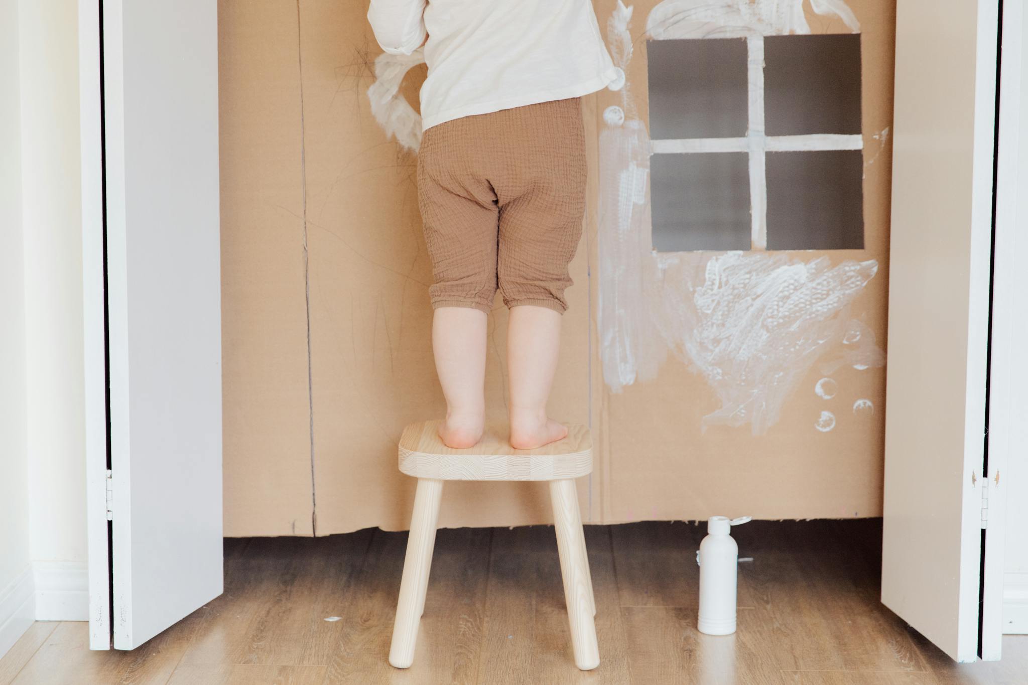 A young child creatively painting on a wall from a wooden stool indoors.